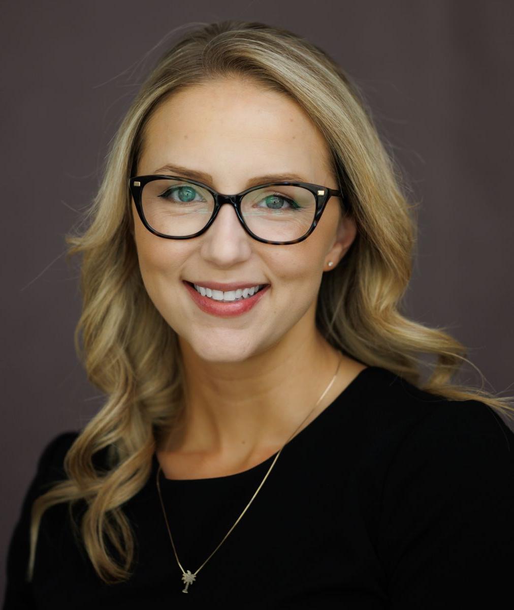 Ms. Marissa Smith, a Caucasian female with long blond hair wears black rimmed glasses and a black suit jacket with a white shirt while smiling at the camera.