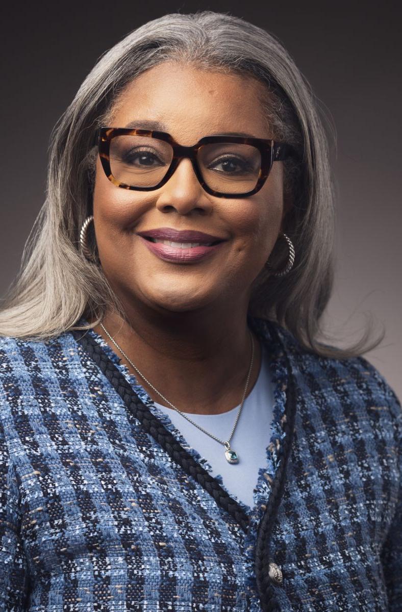 Collette D'Antignac, an African American female, smiles at the camera wearing brown rimmed glasses and a solid blue shirt under a blue plaid sweater.