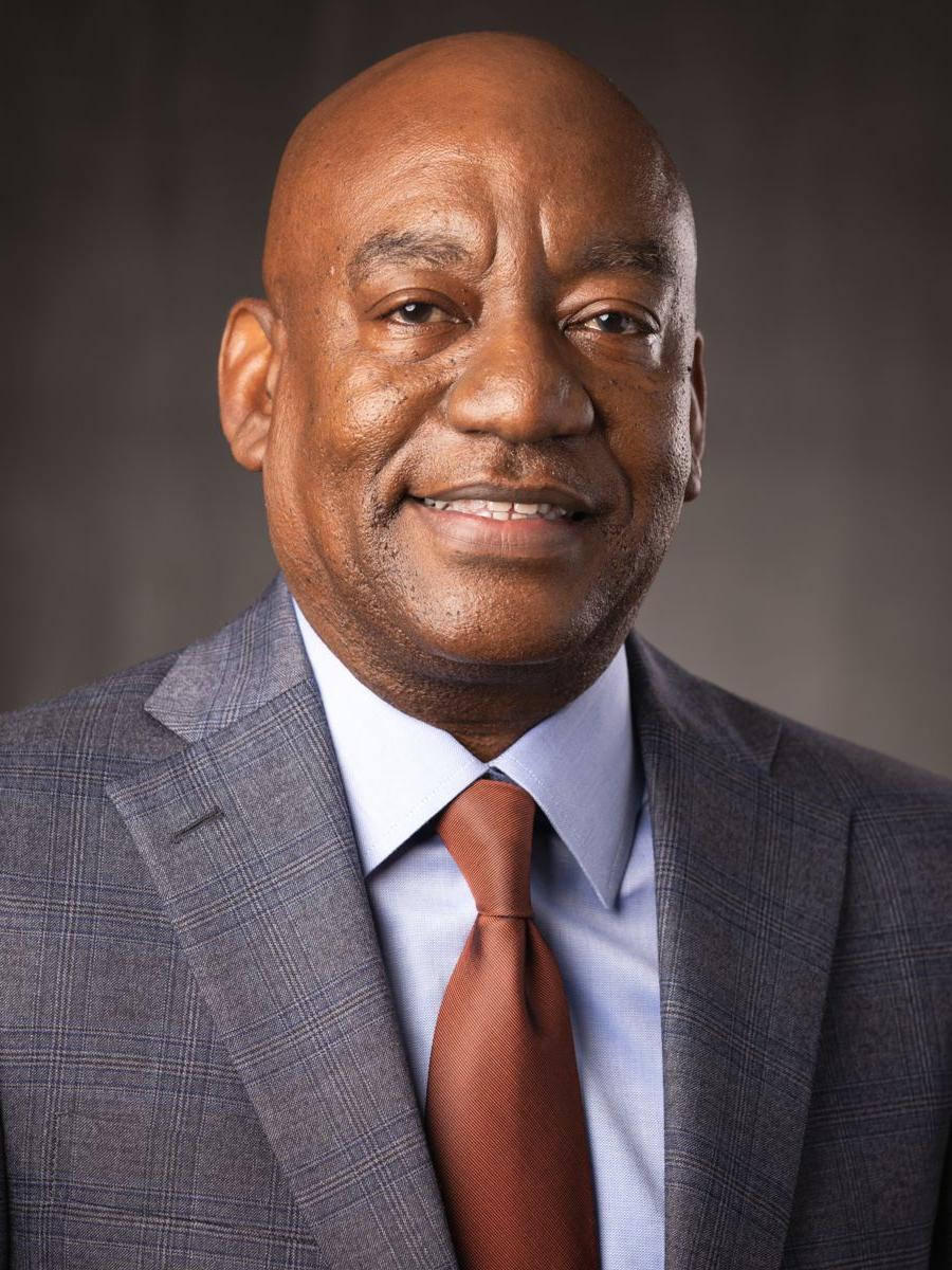 Mr. Derric Terrentine, an African American male, smiles wearing a light blue collared shirt and burnt orange tie with a light grey suit jacket on a grey background.