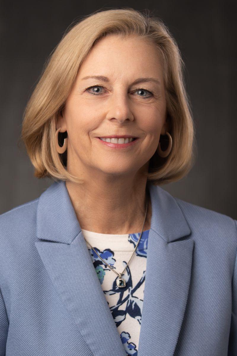 Tammy Shepherd, a Caucasian female with short blonde hair wearing gold earrings,a silver necklace with a blue stone, a white shirt with blue patterns, and a blue suit jacket.